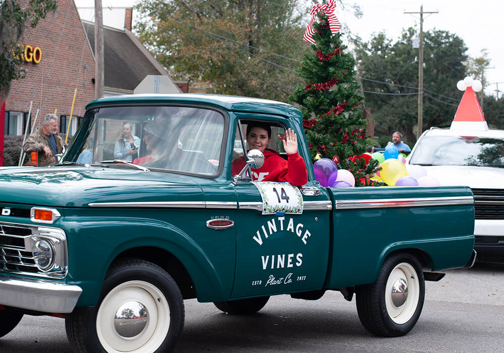 A vintage green pickup truck carries a decorated Christmas tree down the parade route